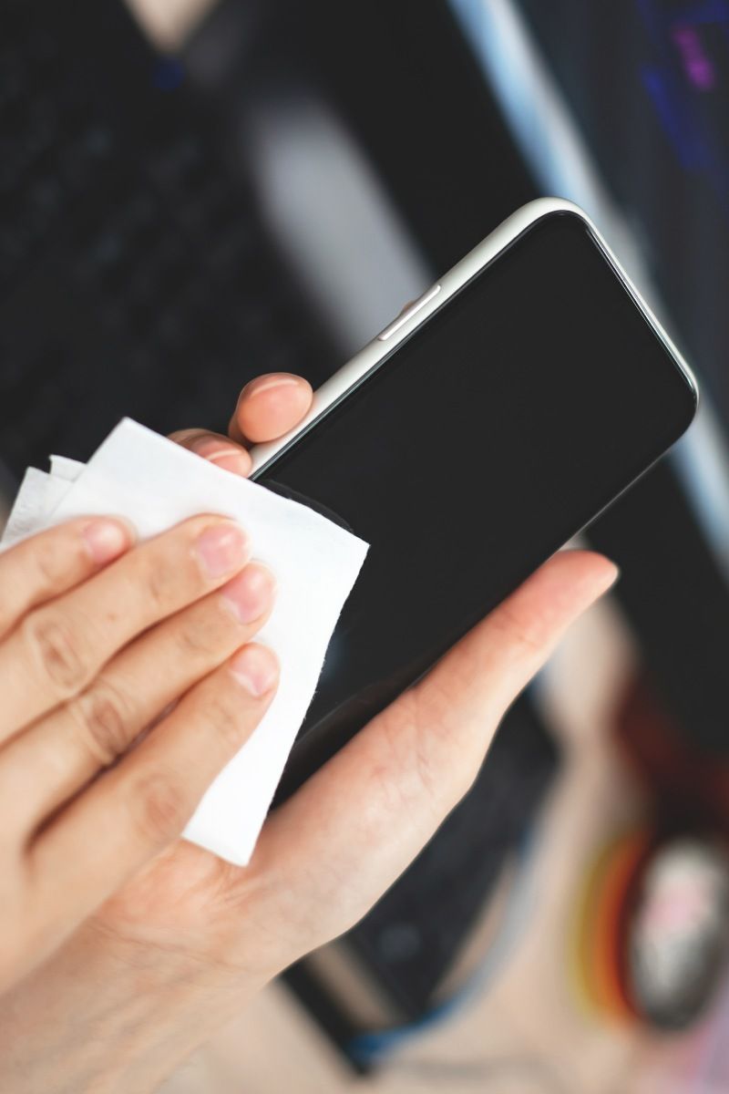 Mano con guante limpiando la parte trasera de un smartphone con un paño humedecido en alcohol.