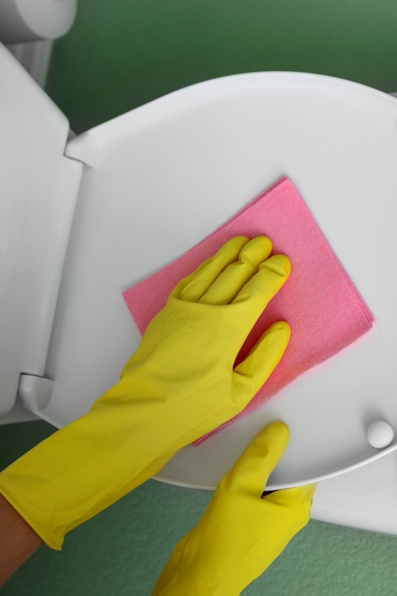 Mano con guante limpiando la tapa de un wc con una balleta.